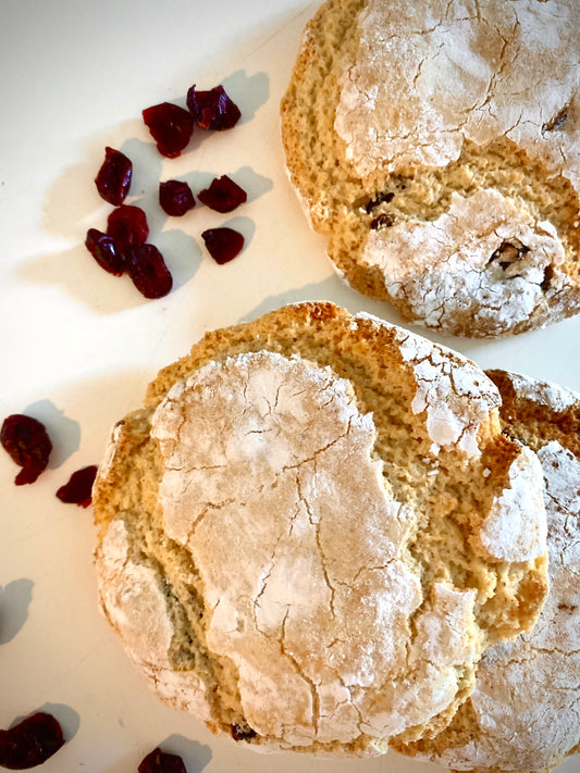 Two golden-brown, rustic-looking vegan cookies with a cracked, textured surface dusted with powdered sugar. A scattering of dried cranberries lies near the cookies, inviting you to indulge in a sweet treat from Sweet Almond bakery.