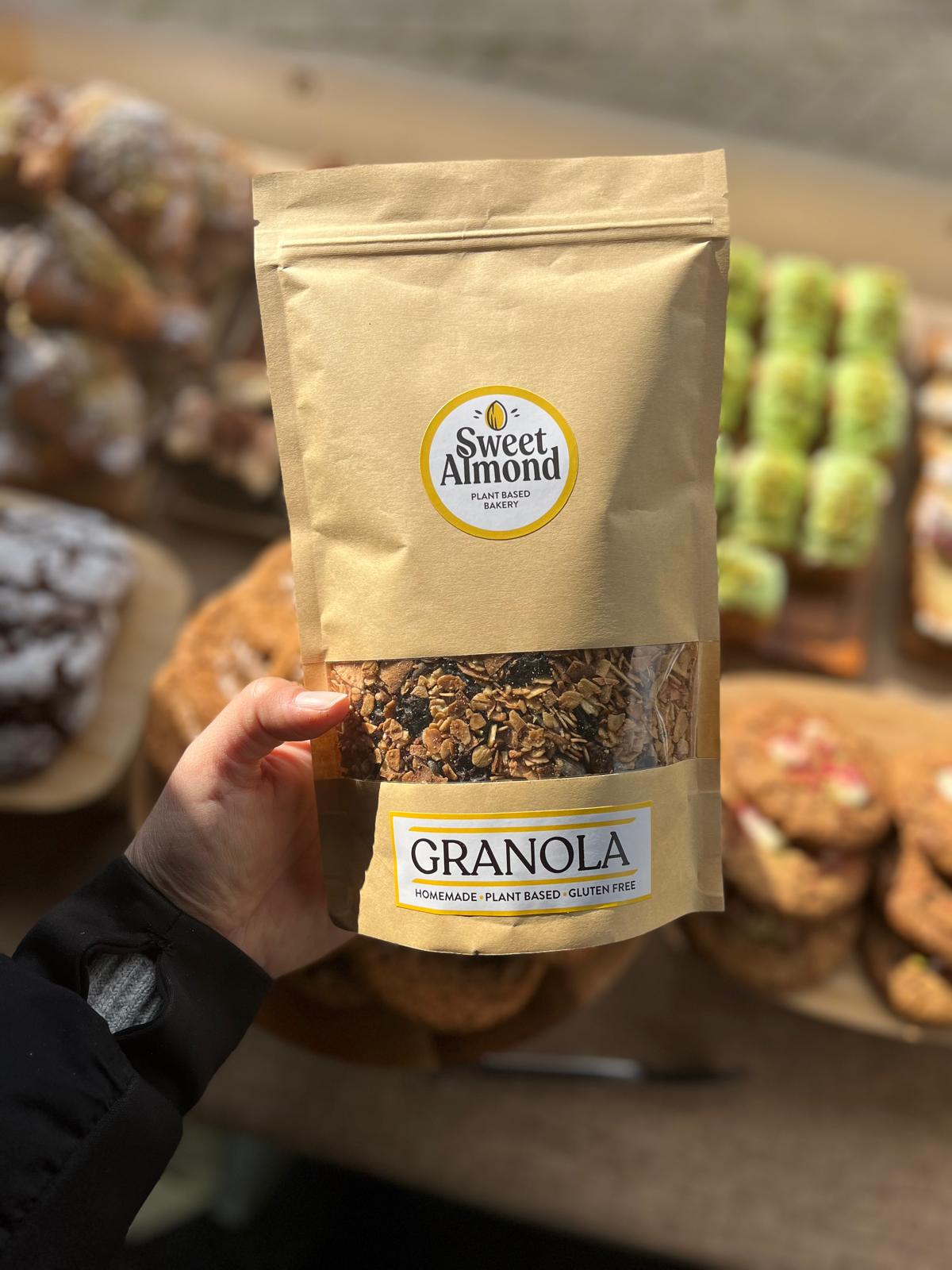 Hand holding a package of Sweet Almond Vegan Granola with pastries in the background