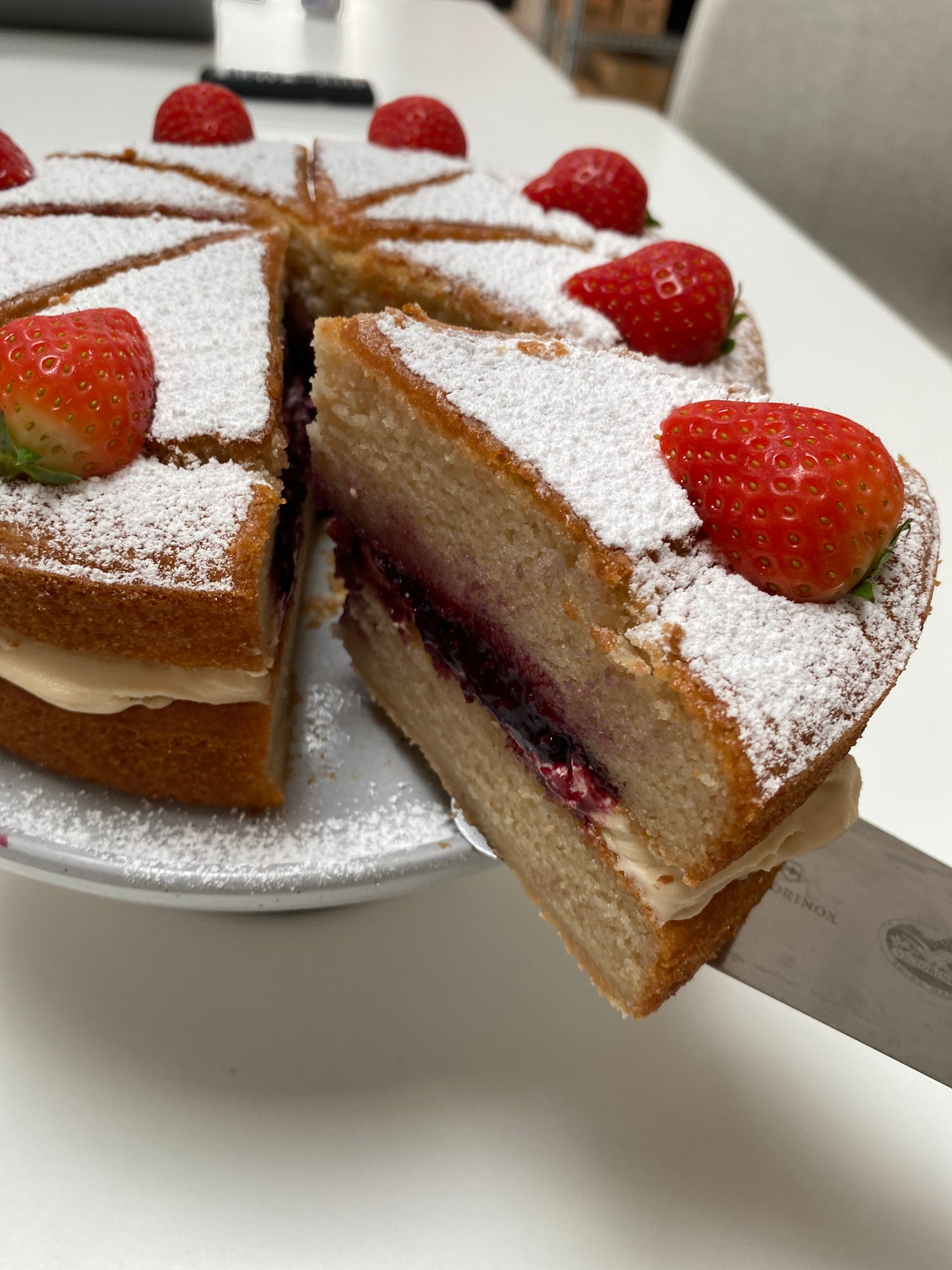 A classic Victoria Sponge Cake, cut in half to reveal a layer of raspberry jam and whipped cream between two fluffy sponge layers, dusted with powdered sugar.